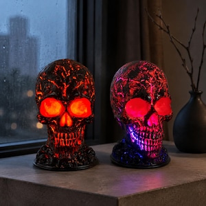 May include: Two skull-shaped lamps with black textured surfaces. The left skull glows with a bright red light from the eye sockets and nasal cavity. The right skull features red light in the eye sockets and a vibrant blue light along the jawline.