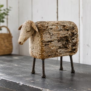 May include: A decorative sheep figurine crafted from a block of weathered, textured wood with a light beige finish. The sheep features a carved head and a rectangular body supported by four thin, dark brown metal legs that stand firmly upright.