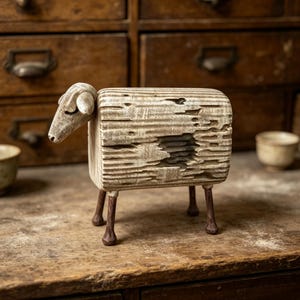 May include: A carved wooden sheep figurine featuring a textured, weathered off-white body with a rectangular block shape, a smooth head with closed eyes, and four thin, dark brown legs. The surface shows deep grooves and a distressed finish.