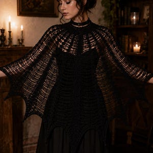 May include: A black crochet capelet featuring an intricate web-like pattern with a high neckline and pointed, bat-wing style hem edges. The garment is constructed with openwork stitching that creates a textured, layered appearance across the shoulders.