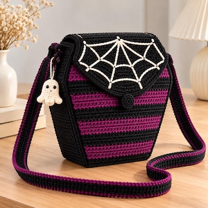 May include: A coffin-shaped crochet crossbody bag featuring horizontal black and purple stripes. The flap displays a white stitched spiderweb design and a small, dangling cream-colored ghost charm attached to the side strap.