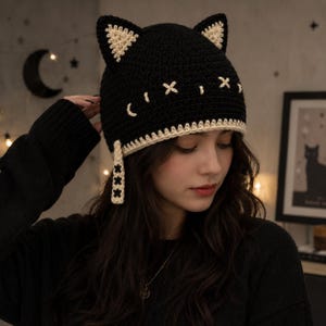 Pode incluir: Um gorro de crochê preto com duas orelhas de gato pontiagudas com detalhes internos em cor creme. O gorro inclui olhos bordados em ponto cruz na cor creme e um acabamento decorativo creme ao longo da borda inferior com uma aba lateral.