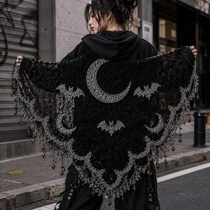 Può includere: Uno scialle triangolare nero con un intricato motivo in pizzo traforato e ricami argentati raffiguranti una grande luna crescente, lune più piccole e pipistrelli. I bordi sono smerlati con perline nere a goccia pendenti.