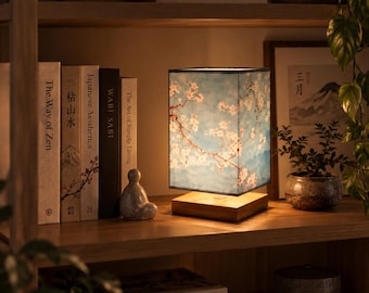 May include: A rectangular table lamp with a light wooden base and a shade featuring a blue background with white cherry blossom branches. The lamp emits a warm glow and is positioned on a wooden shelf next to books and a small gray figurine.