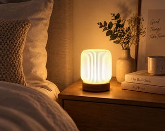 May include: A cylindrical table lamp with a white, vertically ribbed shade and a circular wooden base. The lamp emits a warm, soft glow, illuminating the surrounding area. It sits on a wooden surface next to books and a vase.