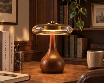 May include: A table lamp featuring a smooth, dark wood base with a teardrop shape and a wide, flat, transparent amber glass shade. The lamp emits a warm, circular glow from an internal light source, positioned on a wooden desk surface.