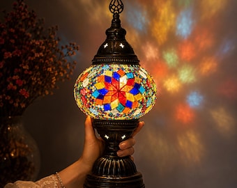 May include: A table lamp featuring a rounded glass shade composed of colorful mosaic tiles in red, blue, yellow, and green geometric patterns. The lamp has a dark metal base and a decorative finial top, casting vibrant light patterns on the wall.