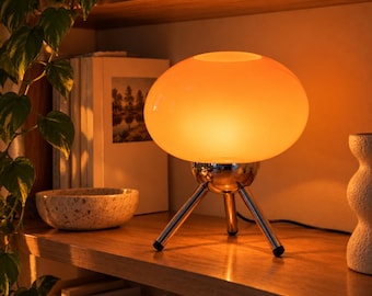 May include: A table lamp featuring a rounded, glowing orange glass shade supported by a polished chrome tripod base with three slender legs. The lamp emits a warm, diffused light, creating a soft illumination across the surrounding area.
