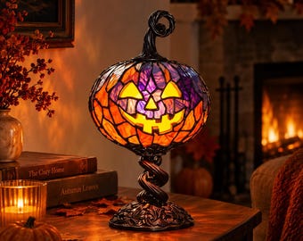 May include: A stained glass jack-o'-lantern lamp featuring orange and purple glass segments with a glowing yellow face. The lamp has a dark, twisted metal stem and a decorative base, emitting a warm light from within the pumpkin-shaped shade.