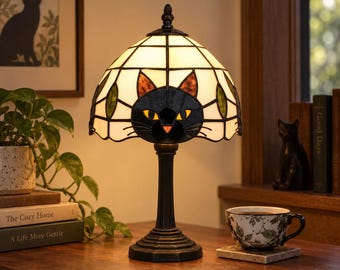 May include: A stained glass table lamp featuring a black cat face design with yellow eyes and orange nose accents. The dome-shaped shade is constructed from white glass panels with green leaf motifs, supported by a dark metal base and a central stem.