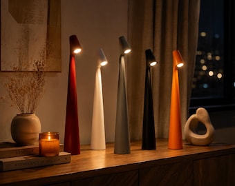 May include: Five tall, slender, conical table lamps in red, white, gray, black, and orange, each featuring a small, angled light source at the top. The lamps are arranged in a row, emitting a warm, focused glow from their individual light heads.