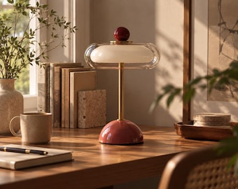May include: A table lamp featuring a rounded, glossy dark red base and a matching dark red sphere finial at the top. The lamp has a slender gold-toned metal stem supporting a horizontal, oval-shaped clear glass shade that encloses a white inner diffuser.