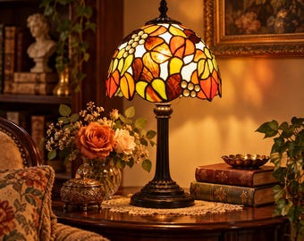 May include: A table lamp with a dome-shaped stained glass shade featuring a pattern of leaves and grapes in shades of orange, yellow, and green. The lamp has a dark metal base with a fluted column and a circular foot, emitting a warm light.