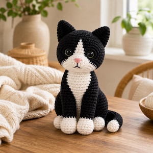 Könnte beinhalten: Ein schwarz-weiß gehäkeltes Katzen-Plüschtier mit weißer Brust, weißen Pfoten und einer kleinen rosa Nase. Die Katze hat große, dunkle, runde Augen und spitze Ohren, einen einfarbig schwarzen Körper und einen weißen Fleck im Gesicht.