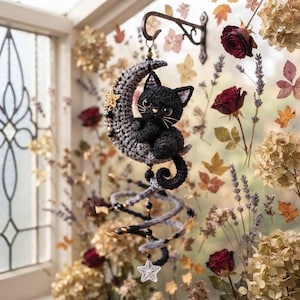 Op de afbeelding: Een gehaakte zwarte kat zittend op een grijze halve maan, opgehangen aan een metalen haak. Het object heeft een spiraalvormige staart, zwarte kralen en een witte ster aan de onderkant. De kat heeft grote donkere ogen en witte snorharen.