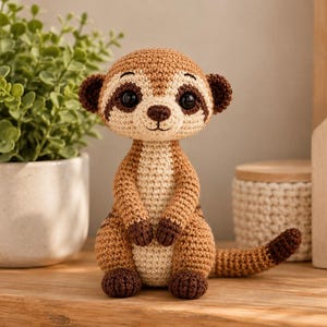 Può includere: Un giocattolo a forma di suricato realizzato all'uncinetto con corpo marrone chiaro, petto e pancia color crema, zampe marrone scuro e macchie marrone scuro attorno ai grandi occhi neri. Il peluche ha piccole orecchie e una lunga coda a strisce.