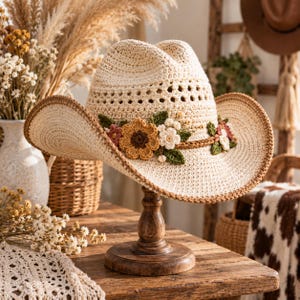 Può includere: Un cappello da cowboy all'uncinetto color crema con una fascia intrecciata marrone decorata da un gruppo di fiori all'uncinetto nei toni del giallo, bianco e rosa tenue, con foglie verdi e un bordo marrone cucito lungo la tesa larga e curva.