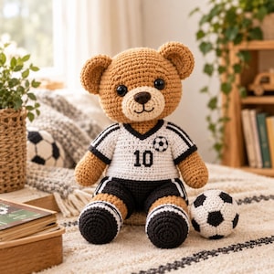 Puede incluir: Un oso de peluche tejido a ganchillo de color marrón que lleva una camiseta de fútbol blanca y negra con el número 10 y un emblema de balón en el pecho, junto con pantalones cortos negros y calcetines, sentado junto a un balón de fútbol tejido.