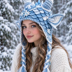 May include: A light blue and white crocheted hat featuring two large horns, pointed ears, and a textured scale pattern. The hat includes snowflake embellishments, long braided side tassels with blue crystal beads, and a thick, warm knit construction.