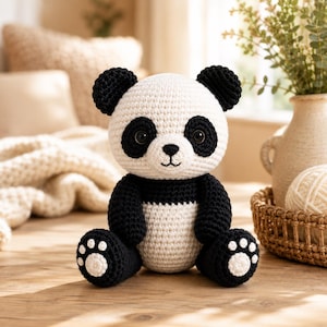 Könnte beinhalten: Ein handgefertigtes Häkel-Panda-Spielzeug mit weißem Körper und Kopf sowie schwarzen Ohren, Augenringen und Gliedmaßen. Der Panda hat eine kleine, schwarz gestickte Nase und einen Mund sowie weiße, kreisförmige Pfotenpolster an den Fußsohlen.