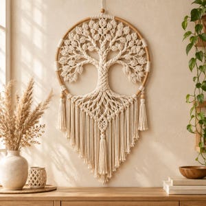 May include: A circular macrame wall hanging featuring an intricate tree of life design crafted from off-white cotton cord. The tree is set within a wooden hoop, accented with two side tassels and a central bottom tassel, creating a textured piece.