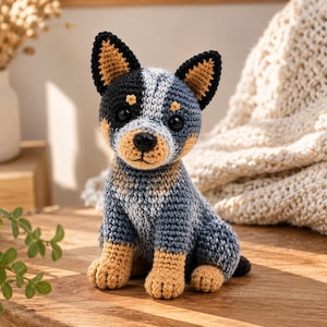 Könnte beinhalten: Ein gehäkeltes Stofftier in Form eines Hundes mit einem Farbmuster in Blau, Grau, Schwarz und Hellbraun. Das Spielzeug hat große schwarze Kunststoffaugen, eine schwarze gestickte Nase, spitze Ohren und eine sitzende Haltung mit Garnstruktur.