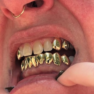 May include: A set of gold teeth grills covering the lower teeth and two upper teeth, with a letter J visible on one of the upper gold caps. The metallic gold finish is highly reflective, showing light highlights across the smooth, curved surfaces.