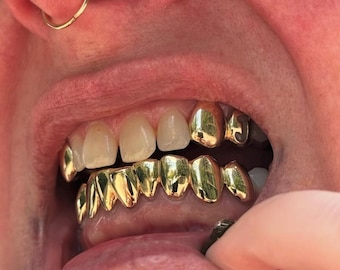 May include: A set of gold teeth grills covering the lower teeth and two upper teeth, with a letter J visible on one of the upper gold caps. The metallic gold finish is highly reflective, showing light highlights across the smooth, curved surfaces.