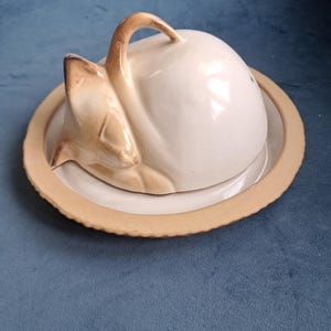 May include: A ceramic butter dish featuring a dome-shaped lid sculpted in the form of a sleeping cat. The cat is off-white with light brown shading on the ears and face. The base is a circular plate with a textured, light brown rim.