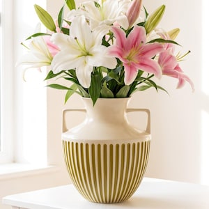 May include: A cream-coloured ceramic vase with two handles and vertical olive green ribbed detailing, filled with a bouquet of white and pink lilies. The text "Pure 1960s Elegance." and "Available now at magpiesnestbyclaire on Etsy" is visible.