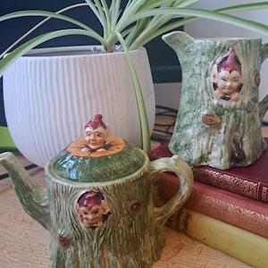 Pode incluir: Um bule de chá e uma leiteira de cerâmica a condizer com um design de tronco de árvore verde texturizado. Ambas as peças são adornadas com pequenas figuras de elfos esculpidas com chapéus vermelhos que espreitam de buracos na casca, esmalte brilhante.