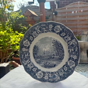 May include: A round ceramic plate with a scalloped edge featuring a dark blue and white transferware pattern. The center depicts a landscape with a building and a boat on water, while the border is decorated with detailed floral motifs.