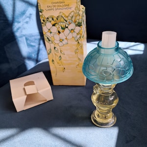 Op de afbeelding: Een Avon Charisma Eau de Cologne miniatuur lampflesje met een doorschijnende blauwe glazen kap met bloemmotief en een heldere glazen voet. De bijbehorende gele kartonnen doos toont bloemillustraties en de tekst Miniature Lamp.