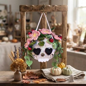 May include: A crocheted skull hanging ornament featuring heart-shaped black eye sockets, a central metallic ring, and two small skull accents. The skull is adorned with pink flowers, green leaves, and small red ladybug embellishments.