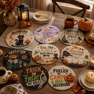 以下が含まれることがあります： Spooky Cat Social Club、Broom Parking Only、The Boo Crew、Trick or Treat Yo' Selfなどのハロウィーンをテーマにしたイラストやテキストが描かれた、8枚の円形装飾コースターのコレクション。木製のテーブルの上に配置されています。