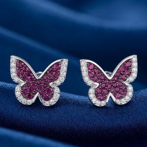 May include: A pair of butterfly-shaped stud earrings featuring a central cluster of small, vibrant magenta-pink stones surrounded by a delicate border of tiny, clear white stones set in a polished silver-toned metal frame.