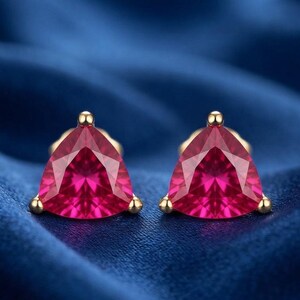May include: A pair of stud earrings featuring vibrant, triangular-cut red gemstones. Each stone is secured by three small, polished gold-tone metal prongs at the corners, creating a clean, symmetrical design that highlights the faceted surface.