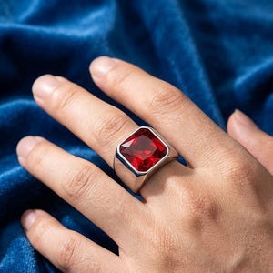 Könnte beinhalten: Ein silberfarbener Siegelring mit einem großen, quadratisch geschliffenen, facettierten tiefroten Edelstein in der Mitte. Die Ringschiene ist glatt und poliert und besteht aus einem breiten, massiven Metall, das sich zum Finger hin verjüngt.