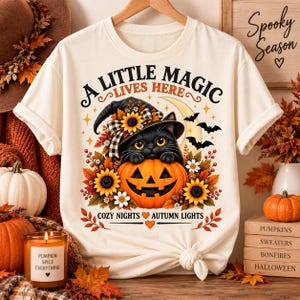 Può includere: Una t-shirt color crema con la grafica di un gatto nero con un cappello da strega, seduto dentro una zucca circondato da girasoli e foglie autunnali. Il testo recita A LITTLE MAGIC LIVES HERE e COZY NIGHTS AUTUMN LIGHTS.