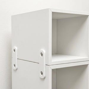 May include: A white modular storage unit with open square compartments. Two white rectangular metal brackets are attached to the side of the unit with silver screws, connecting the stacked sections together to ensure stability and alignment.