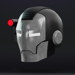 May include: A matte black and metallic silver helmet featuring a rectangular top ridge, glowing white eye slits, and a side-mounted cylindrical attachment with a bright red circular light. The design includes a circular ear piece on the side.