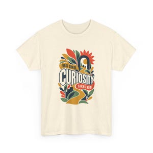 May include: A cream-colored short-sleeve t-shirt with a speckled texture. The center graphic features a winding yellow path, stylized leaves in red, orange, and green, and the text "FIRST GRADE CURIOSITY TAKES ROOT" in a bold, dark blue font.