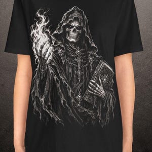 May include: A black t-shirt featuring a detailed white graphic print of a hooded skeleton figure wearing ornate robes and chains. The figure holds a glowing flame in one skeletal hand and a large, heavy-looking book in the other skeletal hand.
