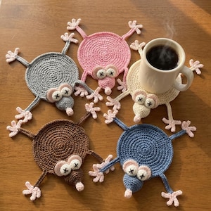 Puede incluir: Cinco posavasos circulares de ganchillo con forma de animales de dibujos animados con cuatro extremidades, un hocico y ojos marcados con una X. Los posavasos son rosas, grises, crema, marrones y azules. Una taza blanca con café está sobre el crema.