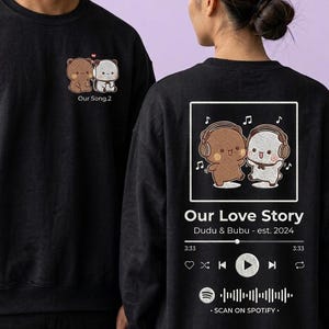 May include: A black sweatshirt featuring a small embroidered graphic of two bears on the chest with the text Our Song.2. The back displays a large graphic of two bears wearing headphones, the text Our Love Story, Dudu & Bubu - est. 2024, and a Spotify-style music player interface with a scannable code.