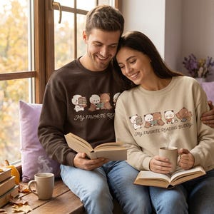 Puede incluir: Una sudadera de cuello redondo color marrón oscuro y una sudadera de cuello redondo color beige, ambas con un gráfico de osos leyendo libros y el texto "MY FAVORITE READING BUDDY" impreso en una fuente cursiva marrón oscuro en el pecho.