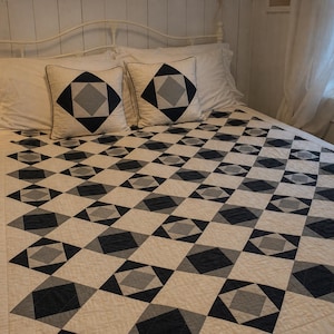 Può includere: Una trapunta e due cuscini quadrati abbinati caratterizzati da un motivo geometrico di rombi color crema, blu navy scuro e grigio chiaro. I pezzi tessili sono trapuntati con un disegno a griglia, mostrando un motivo ripetuto di quadrati e triangoli.