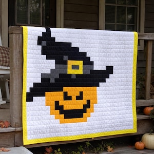 Può includere: Una trapunta quadrata con un design pixelato di una zucca arancione che indossa un cappello da strega nero con una fibbia gialla. La trapunta ha uno sfondo bianco e un bordo giallo brillante, realizzata con cuciture orizzontali e verticali.