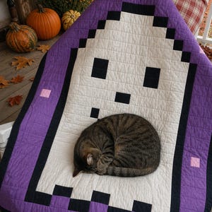 Könnte beinhalten: Eine gesteppte Decke mit einem verpixelten Geisterdesign in Weiß und Schwarz, umrandet von einem breiten violetten Rahmen mit kleinen rosa quadratischen Akzenten. Die Decke ist mit einem gitterartigen Steppmuster über die gesamte Fläche versehen.