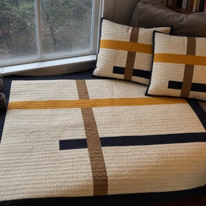 Può includere: Una coperta trapuntata e due cuscini quadrati abbinati con una base color bianco sporco e un motivo geometrico di strisce orizzontali e verticali che si intersecano in giallo senape, marrone e blu navy scuro con texture impunturate.